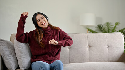 Young cheerful Asian woman listening to music with headphones, dancing on the sofa.