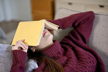 Young Asian woman laying down on the sofa with book cover on her face, taking a nap