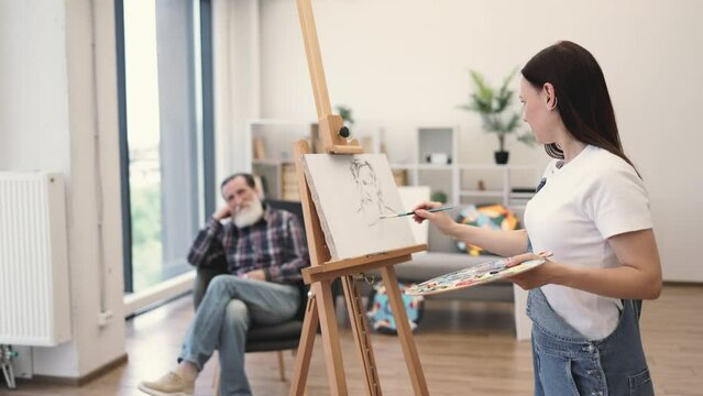 Side view of caucasian lady in cozy clothes applying paint on cotton canvas while keeping eyes on sitter in studio space. Talented amateur artist getting experience by drawing from calm senior man.