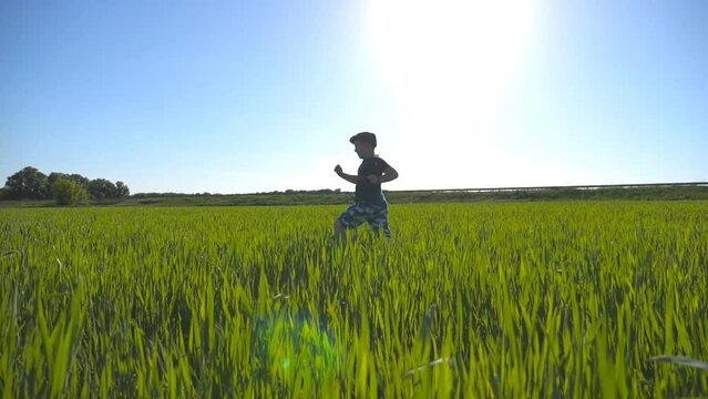 Carefree child in hat jogging at the meadow in summer and having fun. Happy boy is running through the field with green wheat at a sunny hot day. Concept of childhood and rest. Slow motion Side view
