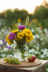 Still life of a bright bouquet of summer flowers against the background of an apple and grapes in the soft setting sun