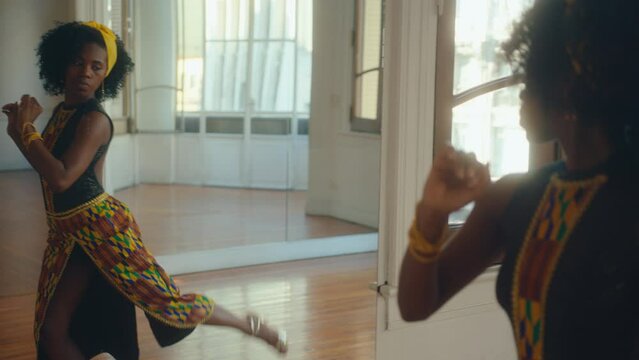 Young inspired African-American female dancer in stage outfit practicing tango movements and steps in front of the mirror during rehearsal in a studio