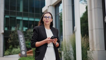 Beautiful confident businesswoman messaging on cell phone typing message browsing internet walking down city street. Woman looking forward and away thoughtfully. Social Networking. Smartphones. Apps. - Powered by Adobe