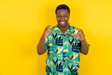 Young latin man wearing hawaiian shirt over yellow background Shouting frustrated with rage, hands...