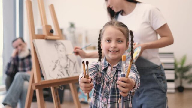 Beautiful girl with paintbrushes looking at camera while young adult drawing from father sitting in background. Talented mother encouraging kid to participate in artistic process at home studio.