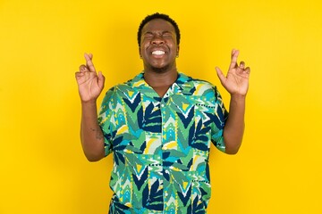 Young latin man wearing hawaiian shirt over yellow background gesturing finger crossed smiling with...