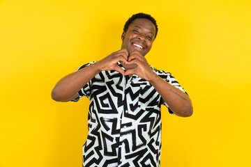 Young latin man wearing printed shirt over yellow background smiling in love doing heart symbol shape with hands. Romantic concept. - Powered by Adobe