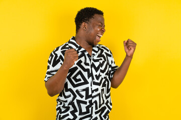Ecstatic Young latin man wearing printed shirt over yellow background shout loud yeah fist up raise...