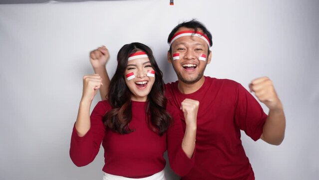 A young Asian couple with a happy successful expression wearing red top and headband while holding Indonesia's flag, isolated by white background. Indonesia's independence day concept.