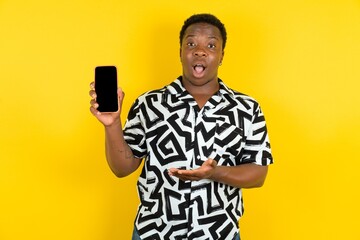 Young latin man wearing printed shirt over yellow background with a mobile. presenting smartphone. Advertisement concept.