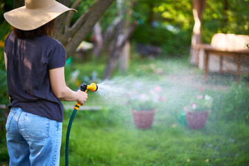 Young woman in hat watering flowers and plants in garden with hose in sunny blooming backyard