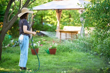 Young woman in hat watering flowers and plants in garden with hose in sunny blooming backyard