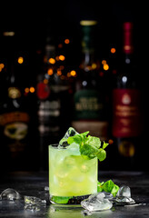 Green Negroni alcoholic cocktail drink with dry gin, aperitif, gentian liqueur, mint and ice cubes. Black bar counter background, bar tools, bottles