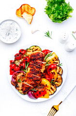 Grilled chicken breast and various vegetables. Colorful paprika, zucchini, eggplant, mushrooms, tomatoes, onion with rosemary on plate, white table background, top view