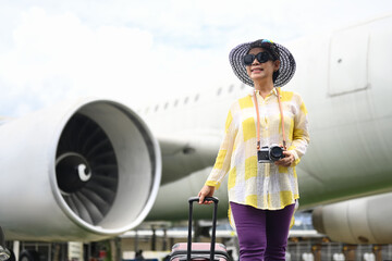 Happy 60s woman in summer clothing and sunglasses walking near the airport with a suitcase.Travel and summer vacation concept