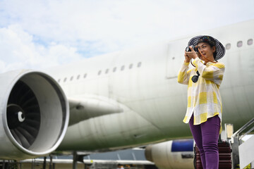 Happy senior woman in casual clothing with hat taking picture at the airport in front of the plane