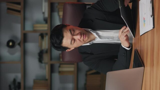 Vertical video of confident asian adult making online call via computer while doing paperwork in office. Intent employee clarifying details of startup project during conversation with colleague.