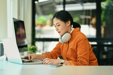 Focused young creative woman with headphone on her neck working on laptop at modern office