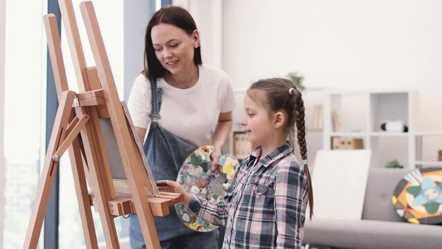 Close up view of involved woman and cute girl applying paint on canvas using brush in studio interior. Cheerful artist teaching playful tween exciting shapes while guiding kid's hand on fabric.