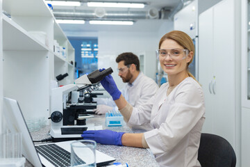 Portrait of female scientist laboratory assistant, researcher working inside medical laboratory with microscope, looking at camera and smiling while sitting, female worker in white medical coat.