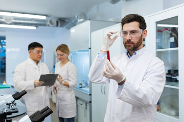 Naklejka na ściany i meble Mature experienced scientist inside laboratory researching and examining blood in test tube, man working with colleagues researchers in white medical coats.