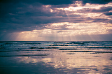 Sunset over Muriwai beach. Sun beams breaking through heavy clouds as waves splashing against the rocks. Auckland, New Zealand