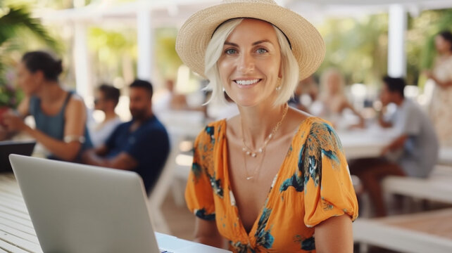 mature adult woman sitting in a cafe or restaurant or club or hotel or holiday resort, with white wooden tables, working on laptop computer, sun hat,
