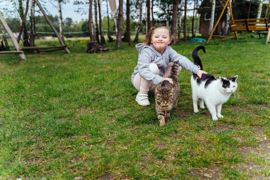 Chereful little girl play with two cats at countryside outdoor. People love pets. A beautiful girl gently hugs and caress her beloved cat while being outside in nature. - Powered by Adobe