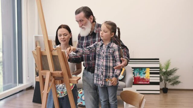 Caring older adult embracing granddaughter near artist easel while young woman fixing paper on canvas. Cute talented girl learning about new textures and shapes during art lesson with mom at home.