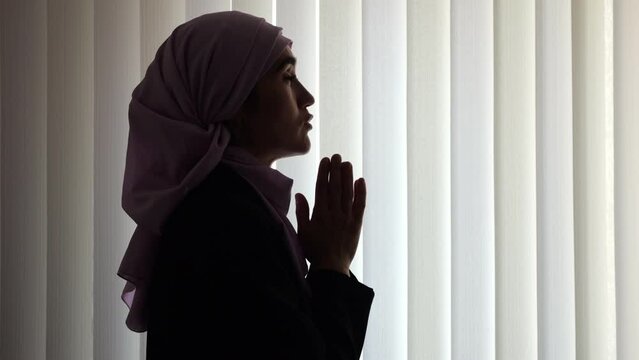 Muslim woman wearing pink hijab prays for more peaceful life in semi-darkened apartment. Religious lady asks Allah for best opportunities for family