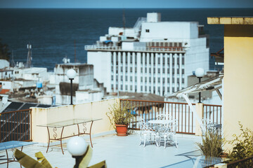 A medium telephone shot with a selective focus on the metal and glass tables on the roof terrace of a hotel house lit by strong tropical sun with the ocean and other houses in a defocused background