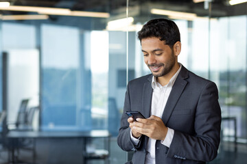 Young arab businessman holding phone, man inside office at workplace typing message and browsing online sites, worker using app on smartphone.