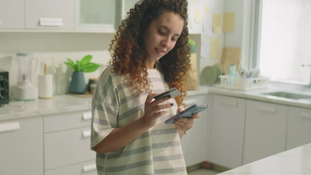 Medium shot of young curly haired girl standing in kitchen at home, using smartphone and credit card while ordering goods from online store