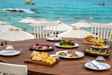 Delicious assortment of dishes (tacos, club sandwich, burger, and snacks) beautifully presented on a table with a mesmerizing sea view in the blurred background. 