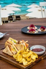 Club sandwich with potato chips, pickles, and ketchup served on a wooden board at a beachfront restaurant with a stunning view of the sea as blurred background. 