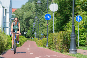 Beautiful girl with sunglasses cycling on the bike road with blue road sign or signal of bicycle lane among green trees and hedges, in spring or summer, nature
