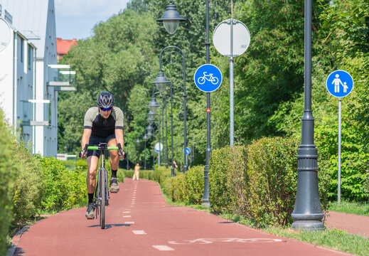 Boy with helmet cycling on the bike road with blue road sign or signal of bicycle lane among green trees and hedges, spring summer nature and street lamps