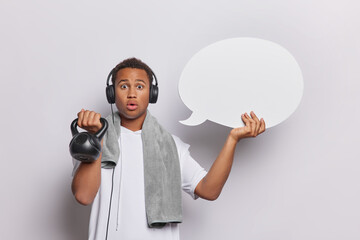 Shocked African man stands impressed does workout at home working on arms strength lifts weight holds blank speech bubble for your advertisement listens music via headphones isolated on white wall