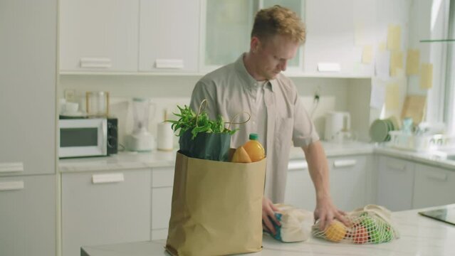 Young man bringing paper bag to kitchen table at home and unloading groceries bought in supermarket