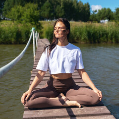 A woman practicing yoga, meditating sitting in a lotus position on a wooden bridge near the lake,...