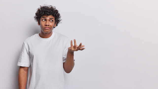 Indoor shot of hesitant Hindu man isolated on white background doubting and shrugging shoulders in questioning gesture looks clueless aside wears casual t shirt blank space aside for your promotion