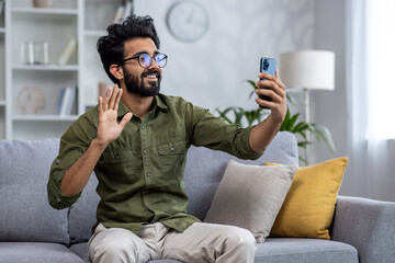 Young smiling man on sofa sitting in living room looking at smartphone camera, waving to friends on online video call and taking selfie photo.