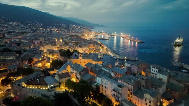 Flying over evening Bastia with city lights, Corsica island. Aerial view of Bastia harbor and old town, ferry leaves the port, mediterranean sea, Corsica, France