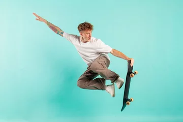 Gordijnen young crazy guy rides skateboard and jumps on blue isolated background, hipster in sunglasses flies © Богдан Маліцький