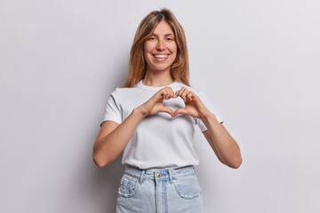 Attractive cute European woman with piercing in nose makes heart gesture expresses love and affection says be my valentine dressed in casual t shirt and jeans isolated over white background.