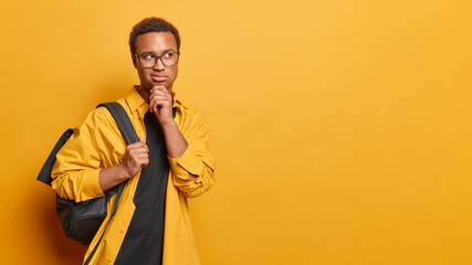 Horizontal shot of thoughtful millennial guy keeps hand on chin concentrated aside pensively considers something wears spectacles and shirt carries rucksack isolated over vivid yellow background