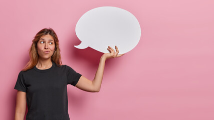Waist up shot of young European woman holds empty speech bubble signaling her intention to share opinion and feedback purses lips dressed in casual black t shirt isolated over pink background