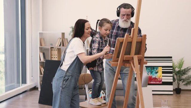 Young barefoot woman with earbuds and mobile watching small girl applying paint on cloth near grandpa with headphones. Creative family practising fine art with musical accompaniment in apartment.