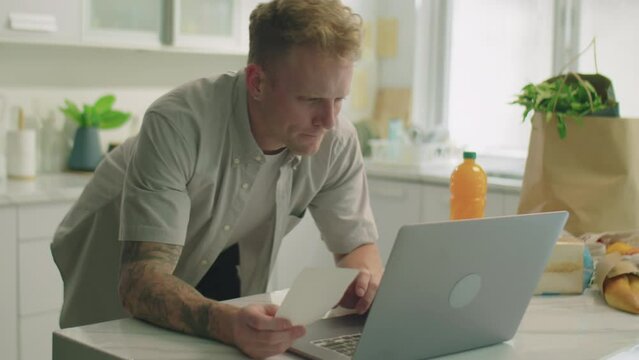 Medium shot of young man leaning on kitchen table, checking receipt and using laptop while calculating cost of groceries and planning budget at home