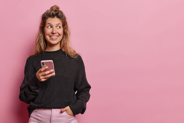 Digital communication concept. Smiling cheerful European woman concentrated aside holds mobile phone browsing new app or website keeps hand in pocket wears casual black jumpet poses indoors.
